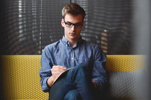 How to practice the Law of Attraction - A young man in glasses writes in a notebook while sitting on a stylish couch indoors.