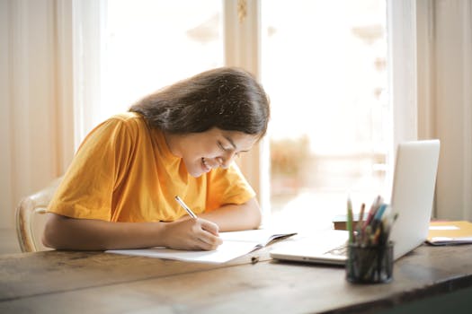 Gratitude and Manifestation - Young woman studying with a laptop and notepad at home, smiling and focused.