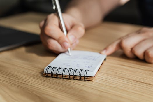 Mindvalley Review - From above of crop faceless male student writing with pen in notebook at wooden desk during lesson