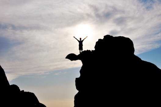 Manifestation Challenges - Silhouette of a person celebrating on a mountain peak against a dramatic sky, symbolizing freedom and success.