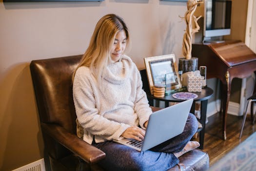 Mindvalley's Duality Review: Is It Suitable For You? Mindvalley Duality Review - Woman seated on a chair using a laptop, enjoying a cozy home office environment.