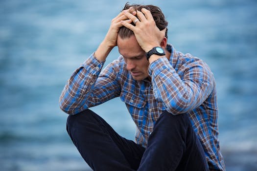 Manifestation Challenges - A man in a plaid shirt sits by the water looking distressed, symbolizing stress.