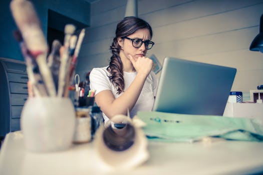 Manifesting Financial Abundance - Young woman with glasses deeply focused on a laptop surrounded by art supplies in a home office.