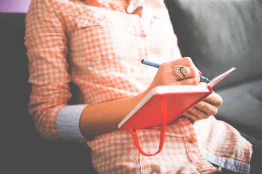 Manifestation Journaling: How to Apply and Key Methods Manifestation Journaling - Close-up of a woman writing in a red journal on a sofa, wearing a stylish ring.
